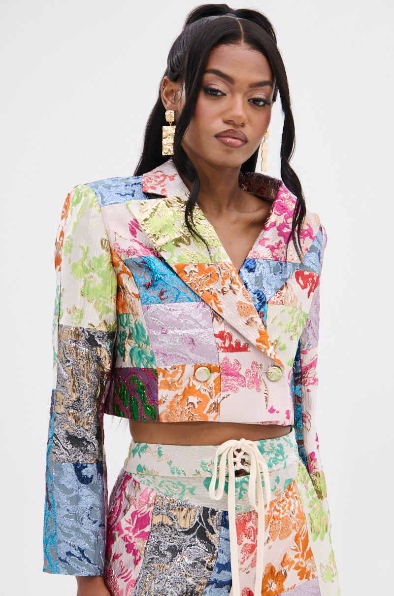 Dressed in the MIXED FEELINGS BROCADE BLAZER and vibrant patchwork pants featuring floral and abstract patterns, a woman with wavy long dark hair showcases long rectangular earrings against a plain background.