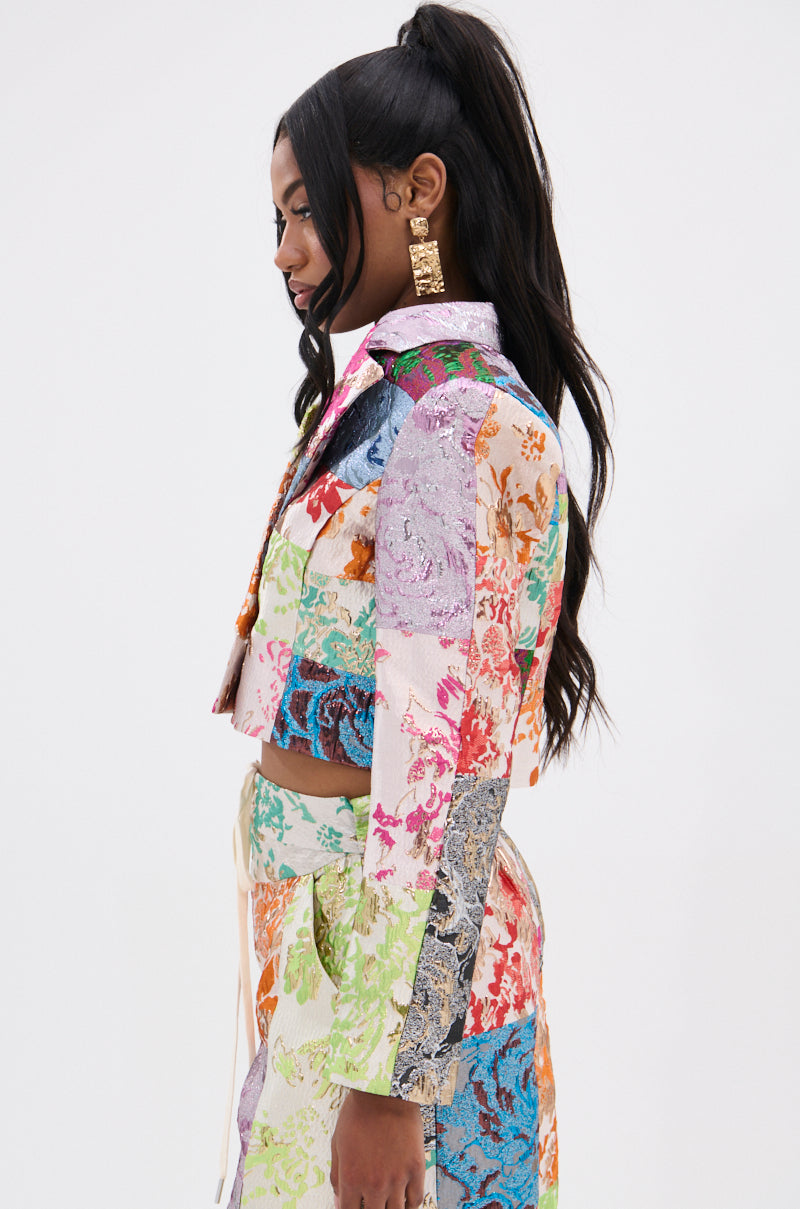 Against a plain backdrop, a woman with long black hair in a high ponytail models the MIXED FEELINGS BROCADE BLAZER and matching skirt, highlighting the vibrant brocade and intricate patchwork design.