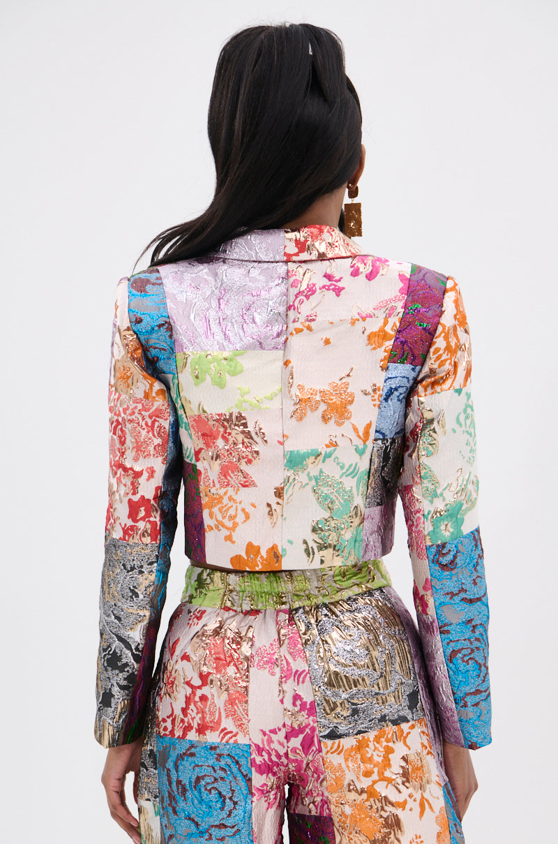 A person with long dark hair wears the vibrant MIXED FEELINGS BROCADE BLAZER and matching pants, featuring a colorful patchwork of floral and textured patterns. They face away from the camera against a plain backdrop.
