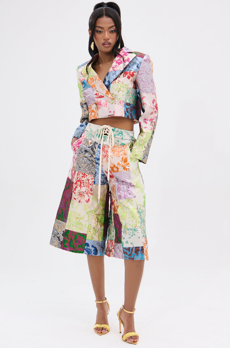 A woman confidently wears the MIXED FEELINGS BROCADE BLAZER and matching knee-length shorts. Her hair is in an updo, and she showcases gold high-heeled sandals. The plain white backdrop highlights her ensemble's intricate patchwork design.