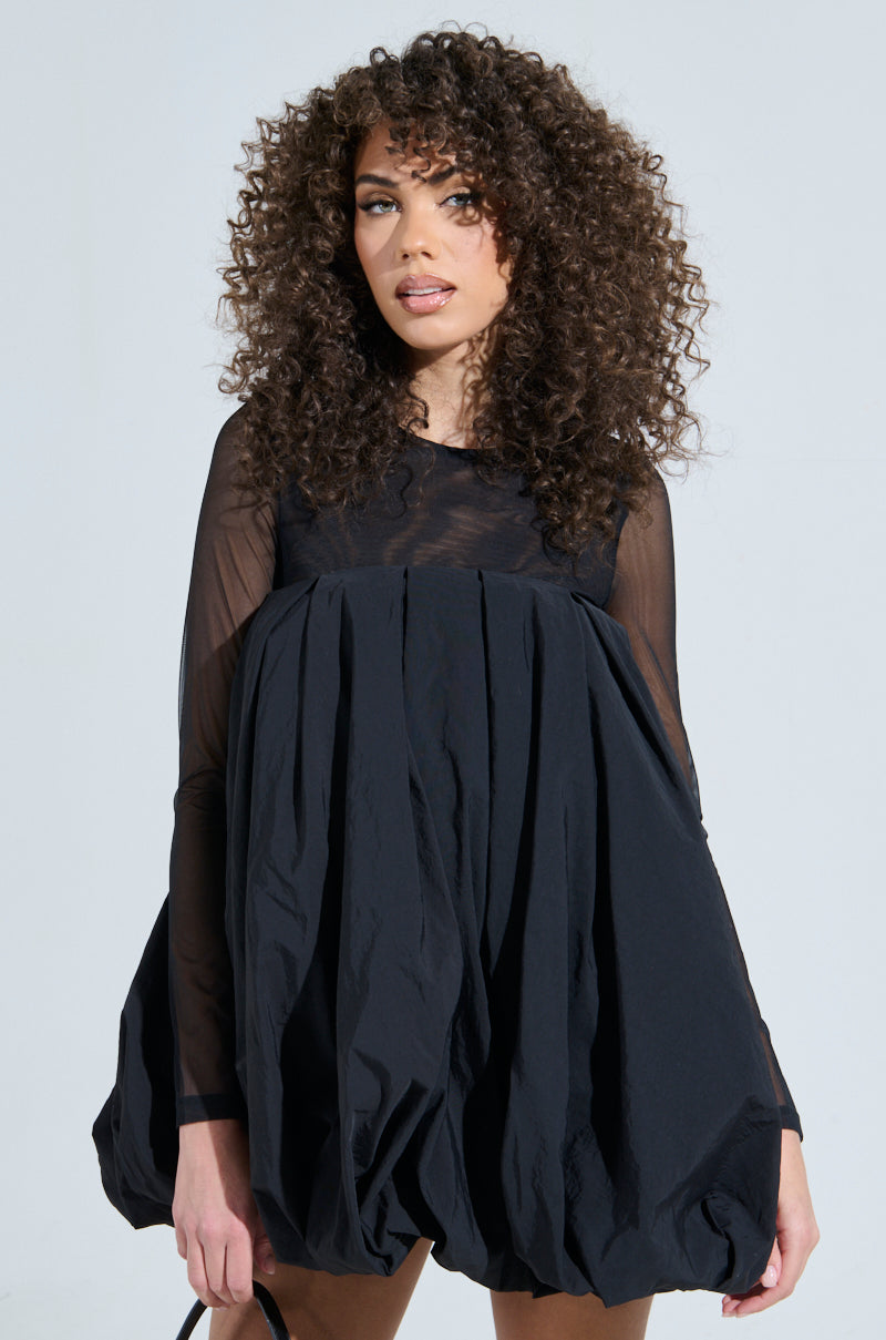 A person with curly hair wears the ISLA BUBBLE MINI DRESS, featuring sheer long sleeves and a voluminous, textured bubble skirt, standing elegantly against a neutral background.