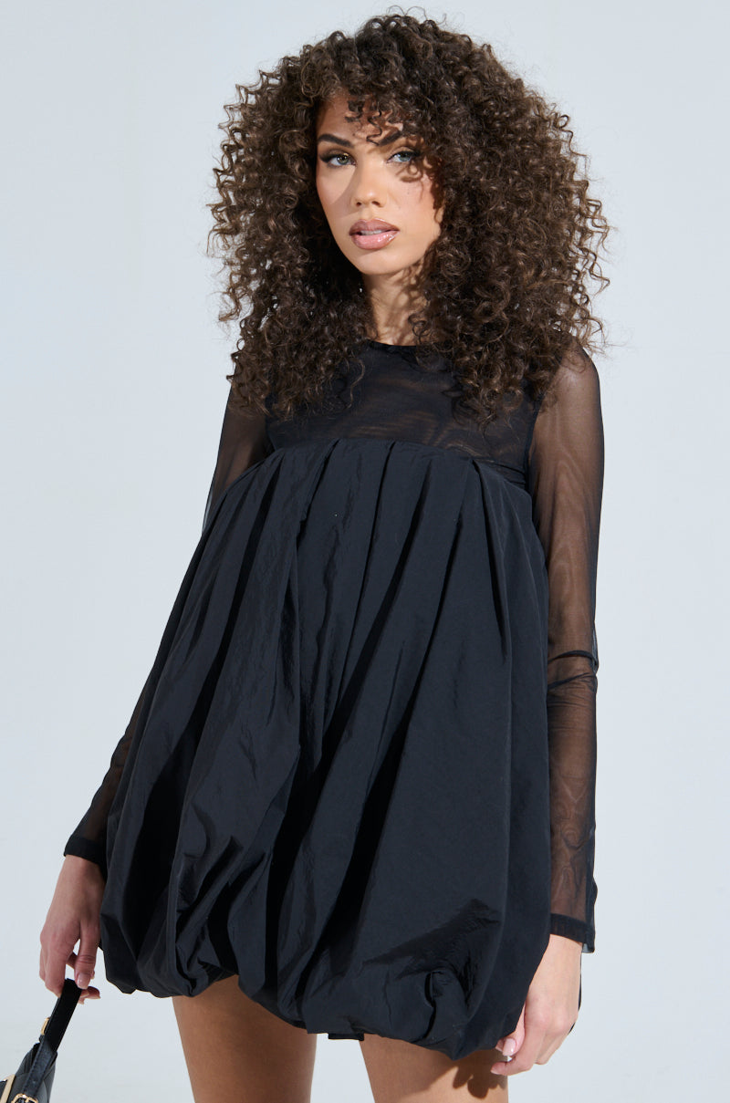 A person with curly hair is wearing the ISLA BUBBLE MINI DRESS, a black long-sleeved outfit with sheer sleeves and a voluminous bubble skirt, set against a plain, light-colored background.