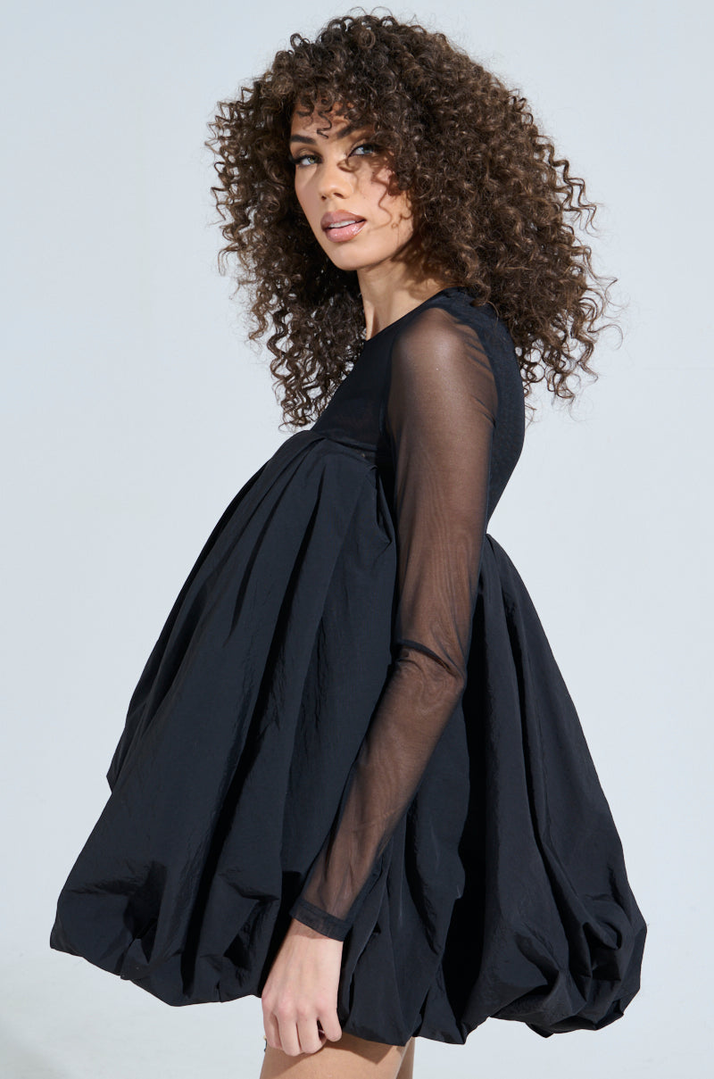 A person with curly hair poses in the ISLA BUBBLE MINI DRESS, a billowy black babydoll silhouette featuring long sheer sleeves and a voluminous bubble skirt. The background is plain and light-colored.