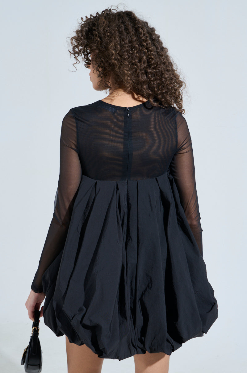 A person with curly hair is wearing an ISLA BUBBLE MINI DRESS featuring sheer sleeves and a pleated skirt in a babydoll silhouette. They carry a small black purse, facing away from the camera against a light background.