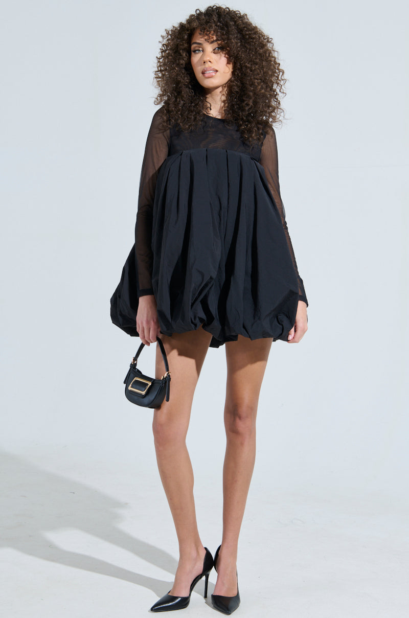 A person with curly hair, wearing an ISLA BUBBLE MINI DRESS and high heels, poses against a plain background. They hold a small black handbag, exuding elegance as they gaze forward.