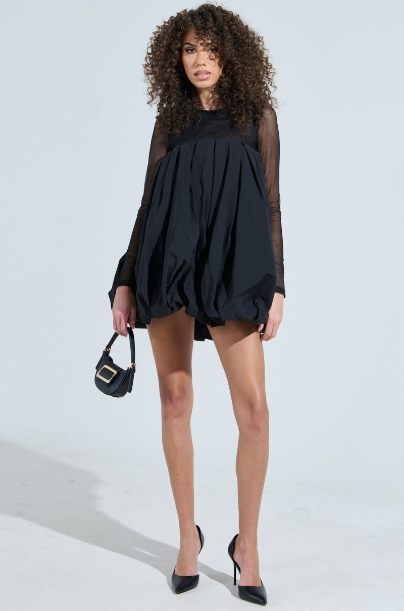 A person with curly hair is confidently posing in an ISLA BUBBLE MINI DRESS featuring sheer long sleeves, holding a small black handbag. They stand against a plain background, wearing black high heels.