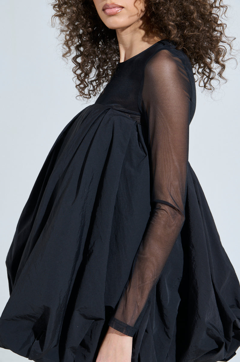 A curly-haired person wears the ISLA BUBBLE MINI DRESS, a loose-fitting LBD with sheer long sleeves and a babydoll silhouette, featuring a voluminous, draped design against a plain light gray background.