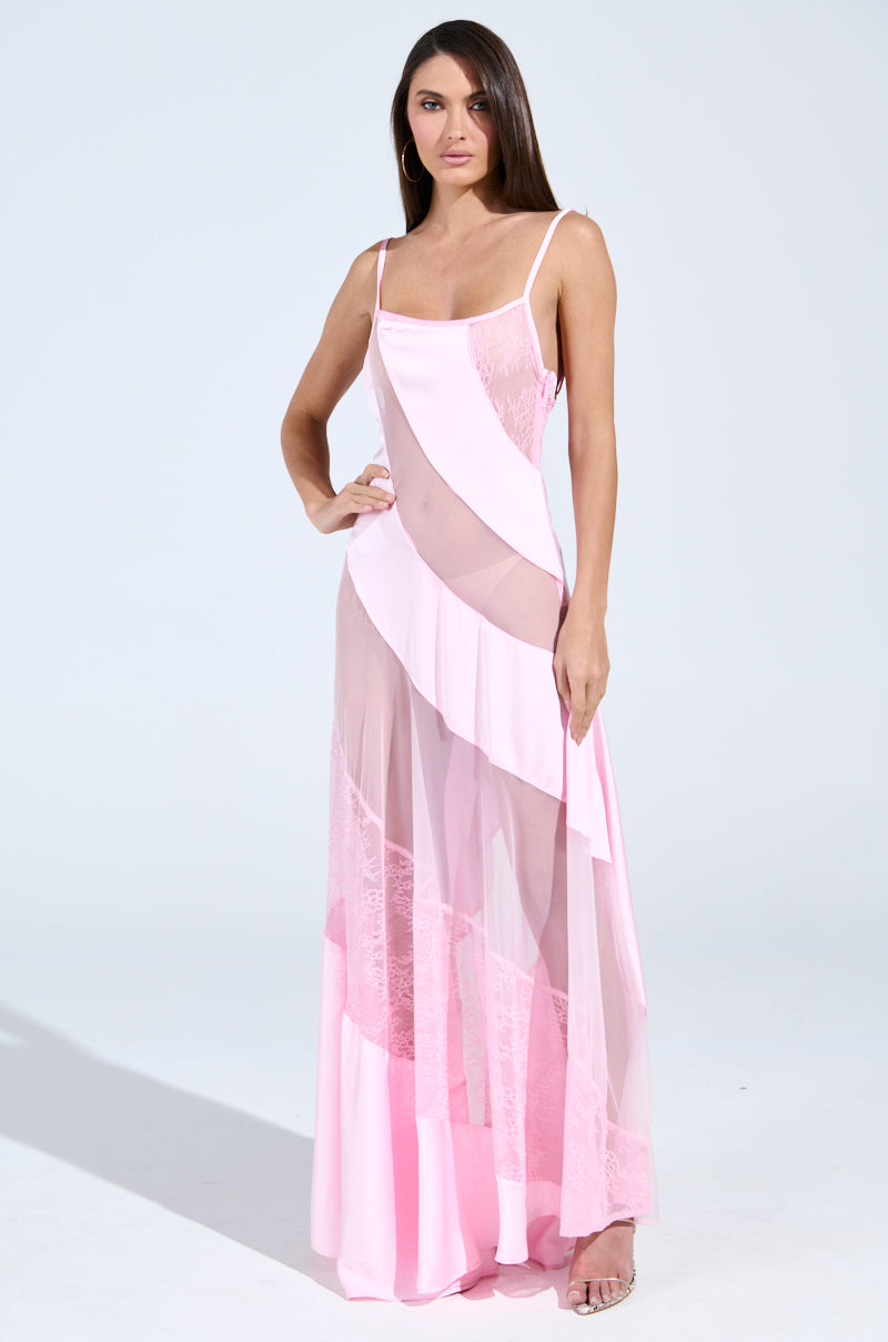 In a sleek pose, a woman dons the ISABELLA SATIN MAXI DRESS, a sleeveless pink gown featuring sheer panels and floral lace details, her hand on her hip against a plain backdrop to highlight its elegance.