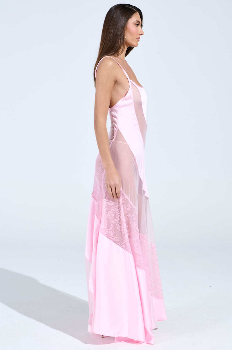A woman models the ISABELLA SATIN MAXI DRESS, highlighting sheer panels and floral lace details, with a sleeveless, floor-length design. The light pink gown's elegant silhouette stands out against a simple light background.