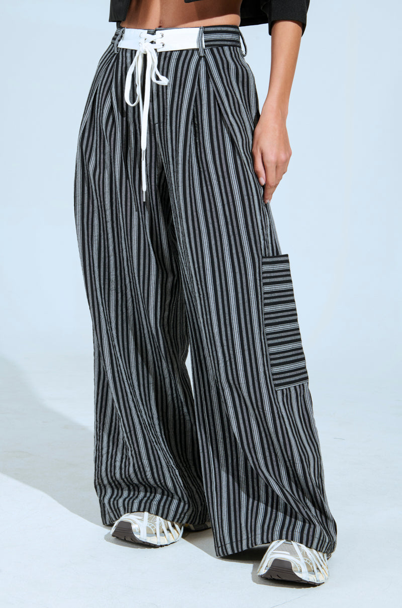 A person wearing the "ALL FALLS DOWN TROUSER," featuring loose, high-rise wide-leg trousers with a black and white striped pattern, a white drawstring waist, and side pockets. The outfit is paired with white and beige sneakers against a plain, light-colored background.