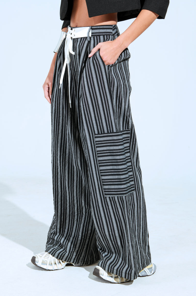Wearing ALL FALLS DOWN TROUSER with a black and white striped pattern and high-rise wide-leg fit, paired with a cropped black top. Their hands are in their pockets and they sport open-toe sandals featuring white strap details against a plain background.