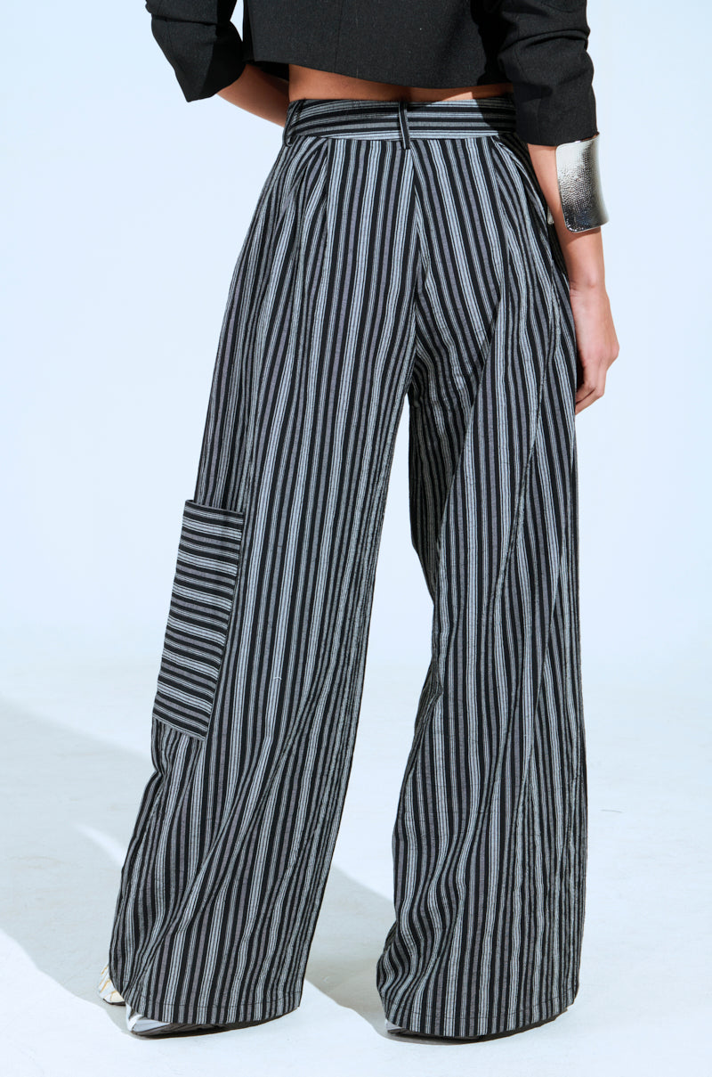 A person wearing ALL FALLS DOWN TROUSER, high-rise with wide-leg and striking black and gray stripes. They feature a large side pocket. Partially visible is a dark top and a metallic bracelet on the wrist.