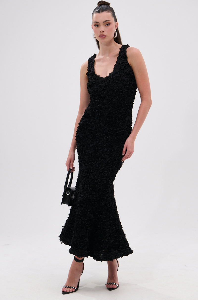 A person wears the GARDEN PARTY FLORAL MIDI DRESS—sleeveless, black, with floral appliqués—paired with a small black handbag and high heels, set against a plain white background.