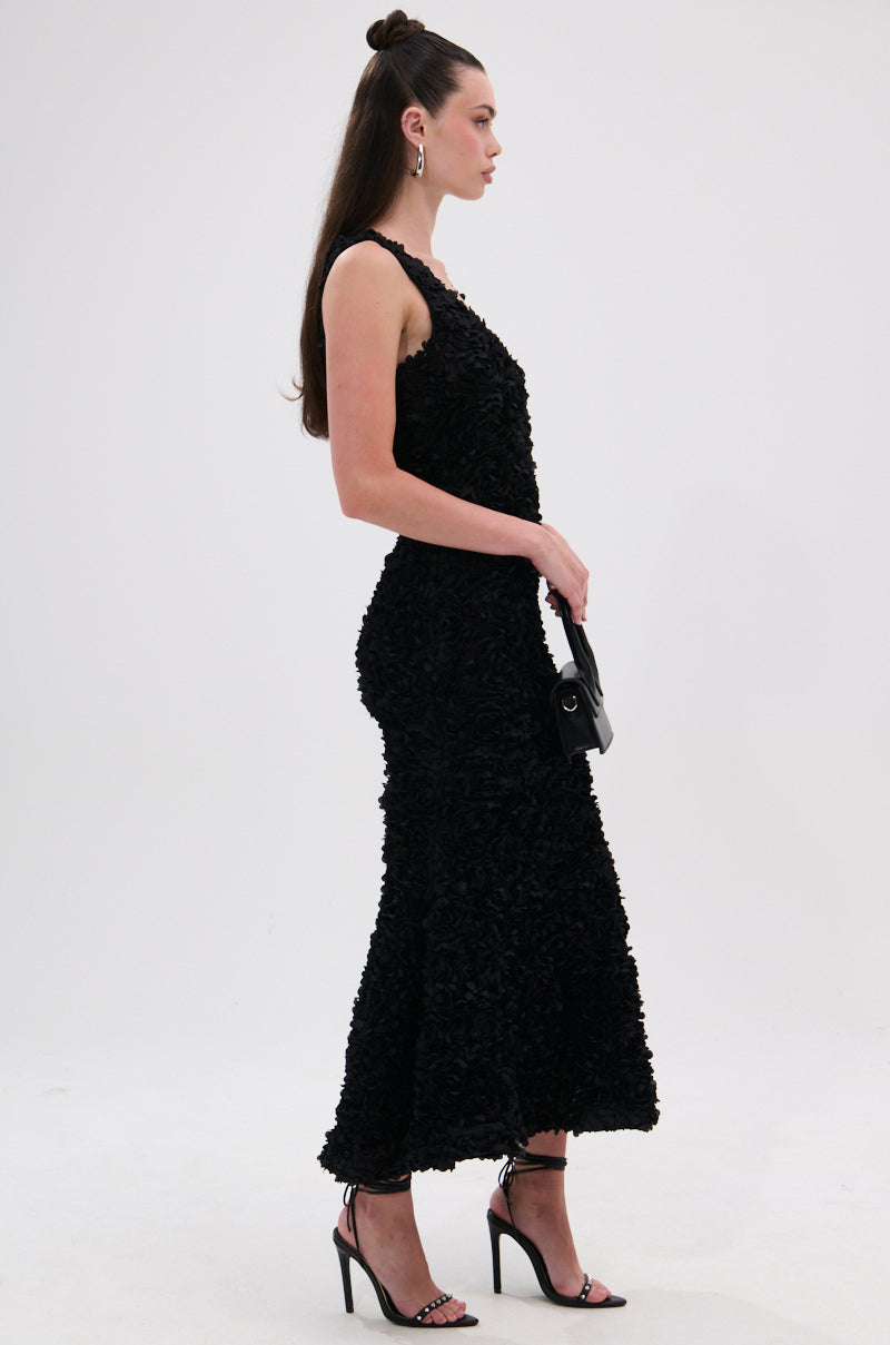 A woman in the GARDEN PARTY FLORAL MIDI DRESS and black high-heeled sandals stands in profile, holding a small black handbag. She has long brown hair styled half-up with hoop earrings against a plain white background.