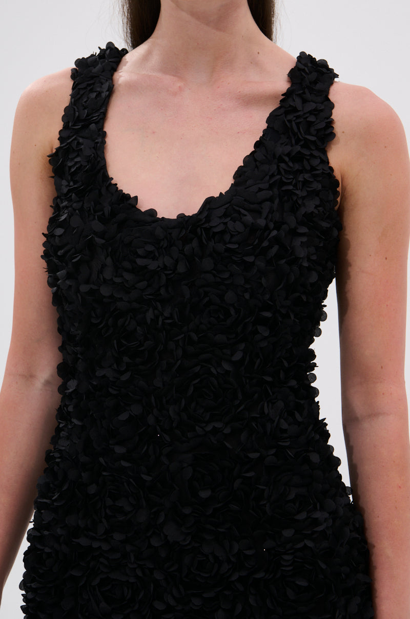 A woman models the GARDEN PARTY FLORAL MIDI DRESS, a sleeveless black dress with textured floral appliqués, visible from shoulders to mid-torso against a plain light background.