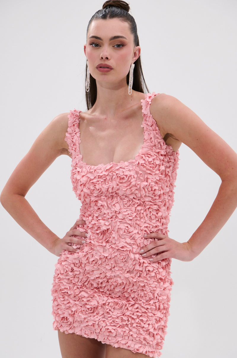A woman stands confidently with her hands on her hips, wearing the LEXI FLORAL MINI DRESS—a sleeveless, curve-hugging mini in light pink, detailed with textured 3D floral appliqués—set against a plain white background. 1