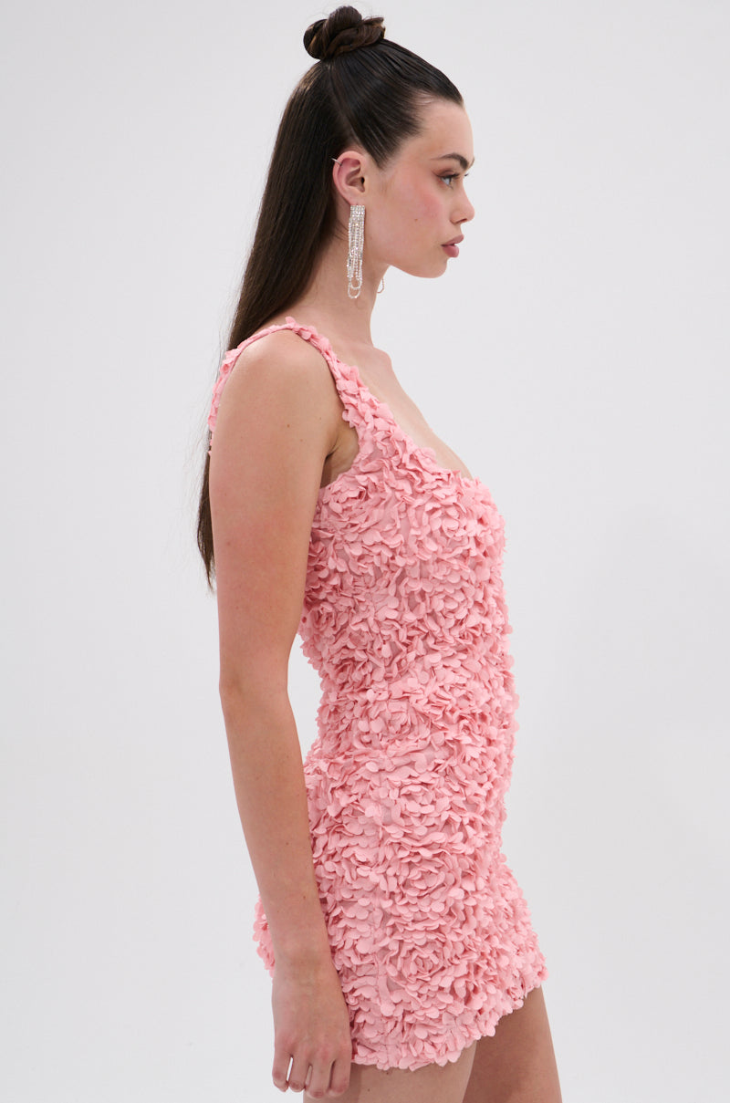 A woman in profile models the LEXI FLORAL MINI DRESS—a sleeveless, curve-hugging pink dress with 3D floral appliqués. Her long brown hair is half up and she wears dangling earrings, set against a plain light background. 2