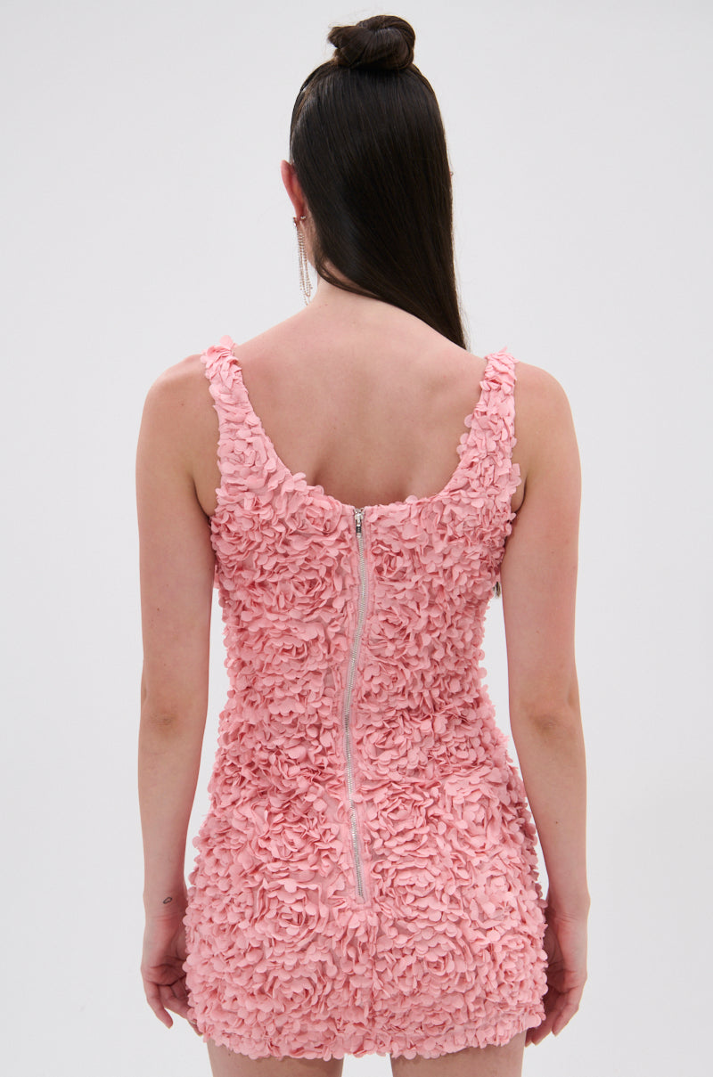 A woman with straight, dark brown hair in a half-up bun stands with her back to the camera, wearing the LEXI FLORAL MINI DRESS—a light pink mini dress with a curve-hugging fit and visible back zipper—against a plain light background. 3
