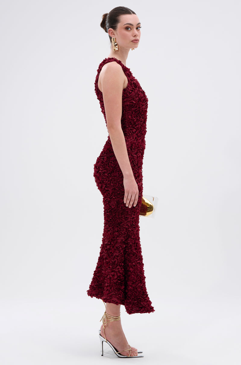 A woman in the GARDEN PARTY FLORAL MIDI DRESS stands sideways, holding a small gold clutch. She pairs it with gold drop earrings, white high-heeled sandals with gold ankle straps, and her hair styled in a sleek bun.