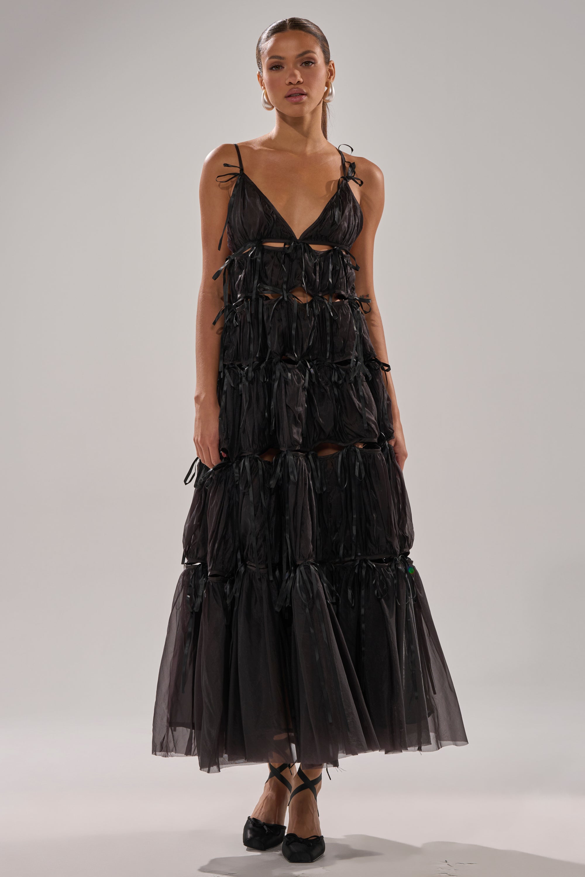 A woman wears the COULDN'T BE ME IF YOU TRIED TIE MAXI DRESS IN BLACK, a long tiered chiffon dress with cut-out details and thin shoulder straps, paired with black ballet flats against a plain neutral background.