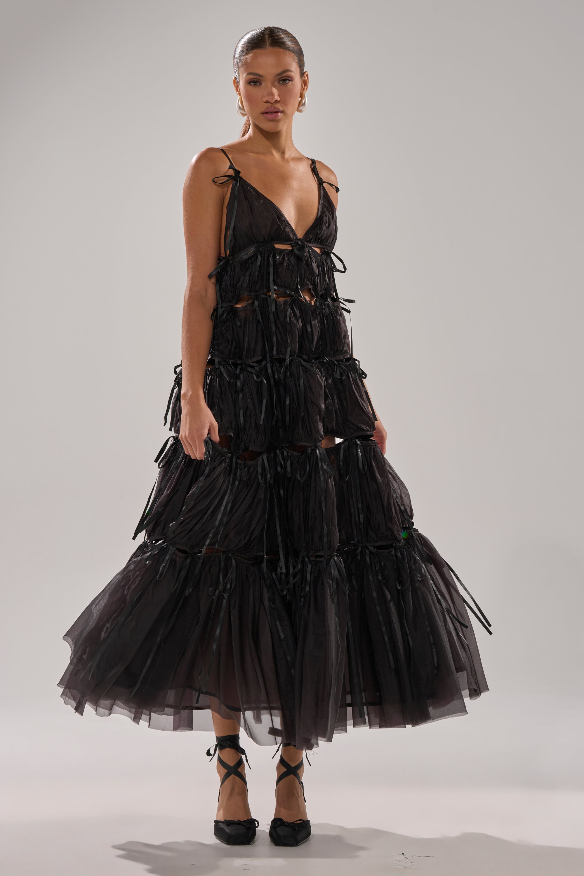 A woman stands confidently against a gray background in the COULDN'T BE ME IF YOU TRIED TIE MAXI DRESS IN BLACK—tiered, sheer tulle with ribbon details and spaghetti straps—paired with black lace-up shoes.