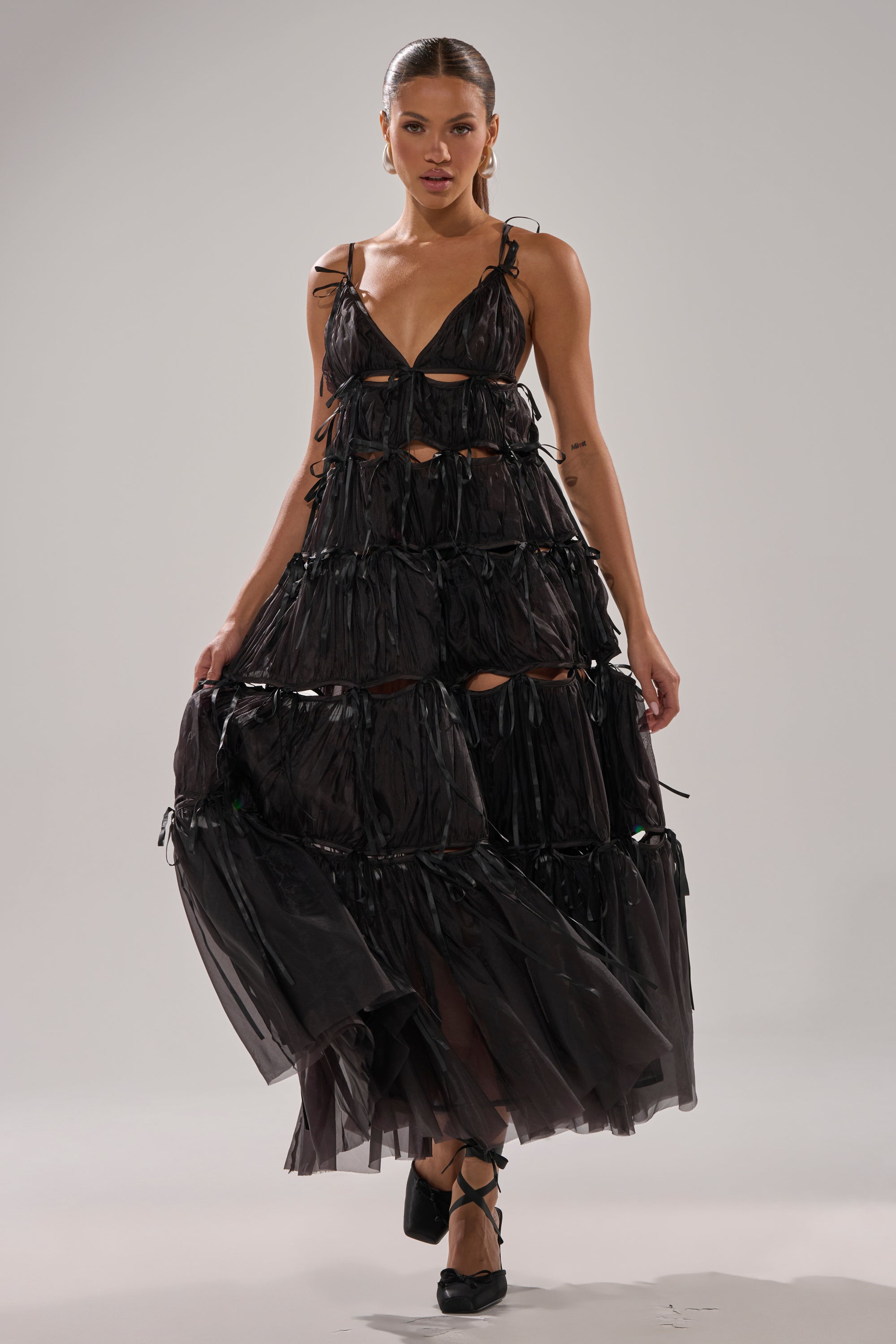 A woman poses in the COULDN'T BE ME IF YOU TRIED TIE MAXI DRESS IN BLACK, featuring tiered strappy cutouts and a flowing chiffon skirt, paired with black lace-up shoes, against a plain light gray background.