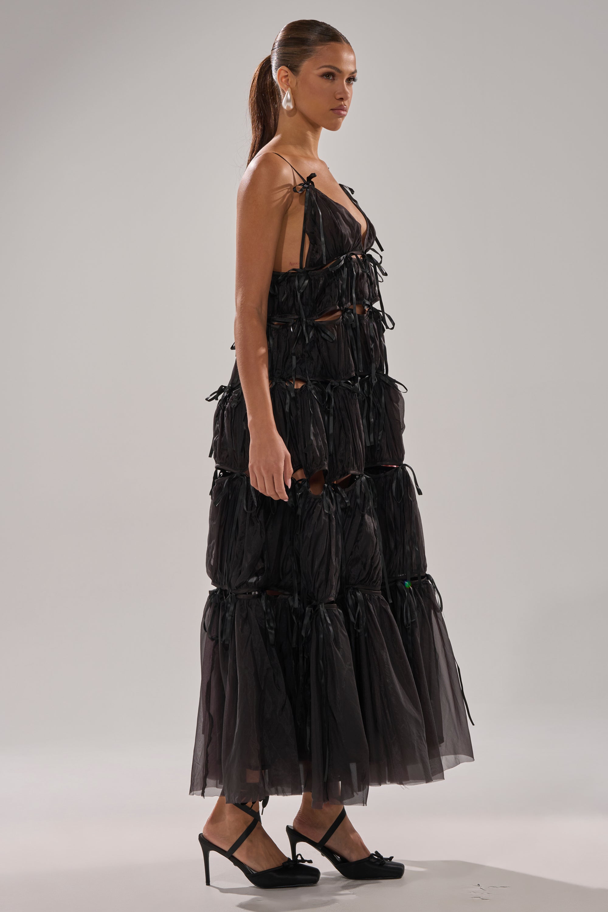 A woman with a ponytail wears the COULDN'T BE ME IF YOU TRIED TIE MAXI DRESS IN BLACK—a sleeveless, tiered chiffon dress with bows—styled with heels and statement earrings, standing against a plain light gray background.