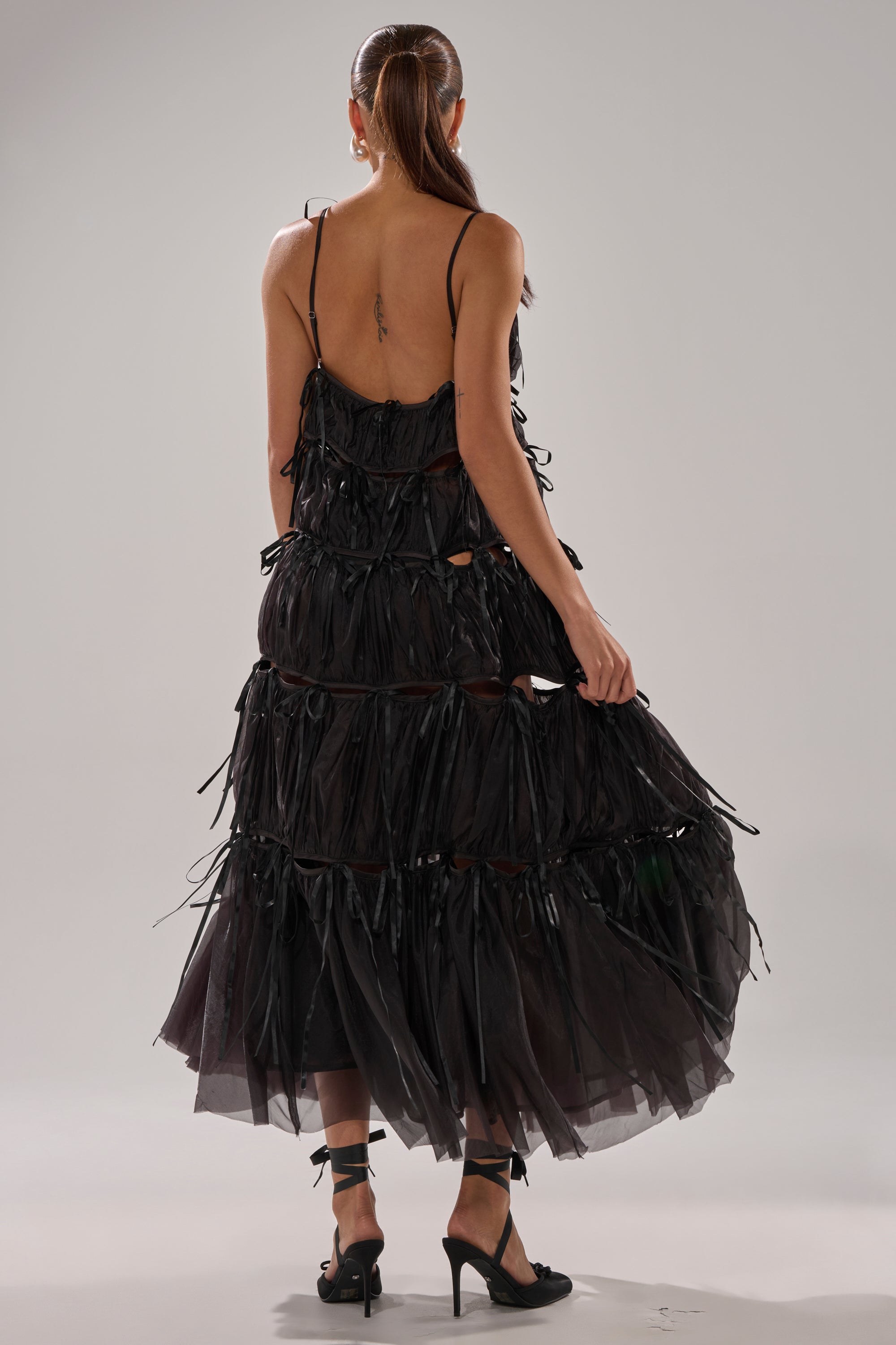 A woman with a ponytail, facing away, wears the COULDN'T BE ME IF YOU TRIED TIE MAXI DRESS IN BLACK with ribbon details and an open back, paired with black heels. She holds out one side of the skirt against a plain light gray background.