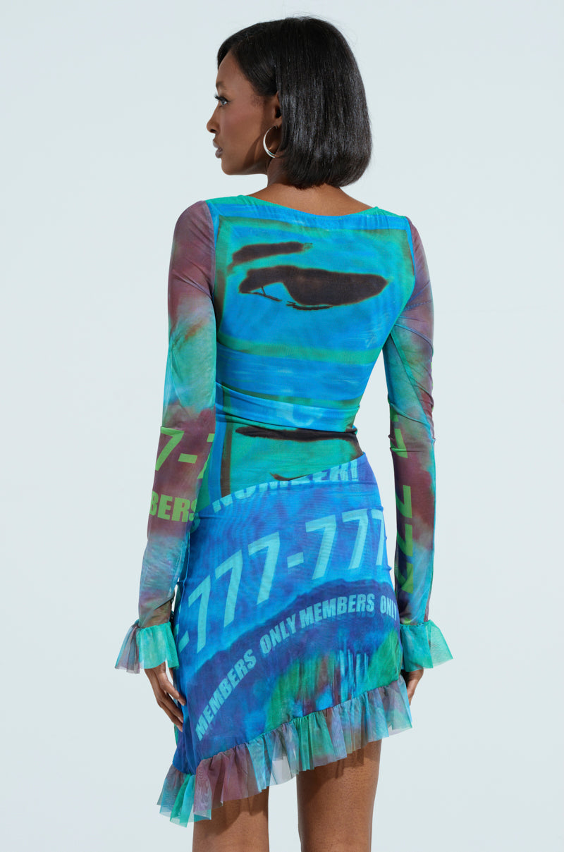 A woman in an AKIRA Label MEMBERS ONLY PRINTED MINI DRESS exudes elegance with its blue and green hues. The ruffled mini dress, adorned with "777-777," features a fitted top and playful hem. She accessorizes with hoop earrings, her hair cascading as she gazes away from the camera.