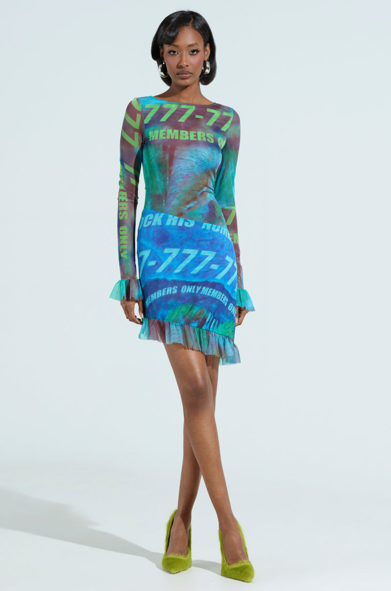 A woman confidently wears a colorful MEMBERS ONLY PRINTED MINI DRESS featuring text and graphic patterns in blue, green, and purple shades. She pairs it with green furry heels, with her hands gently resting in front.