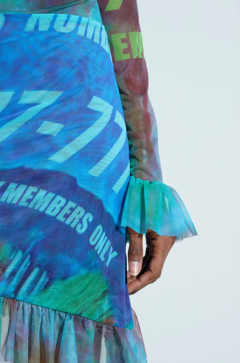 A close-up shows someone in an AKIRA Label MEMBERS ONLY PRINTED MINI DRESS, featuring long sleeves with ruffled cuffs. The blue and green tie-dye fabric displays "NUMBERS" and "MEMBERS ONLY," set against a light blue background for a captivating look.