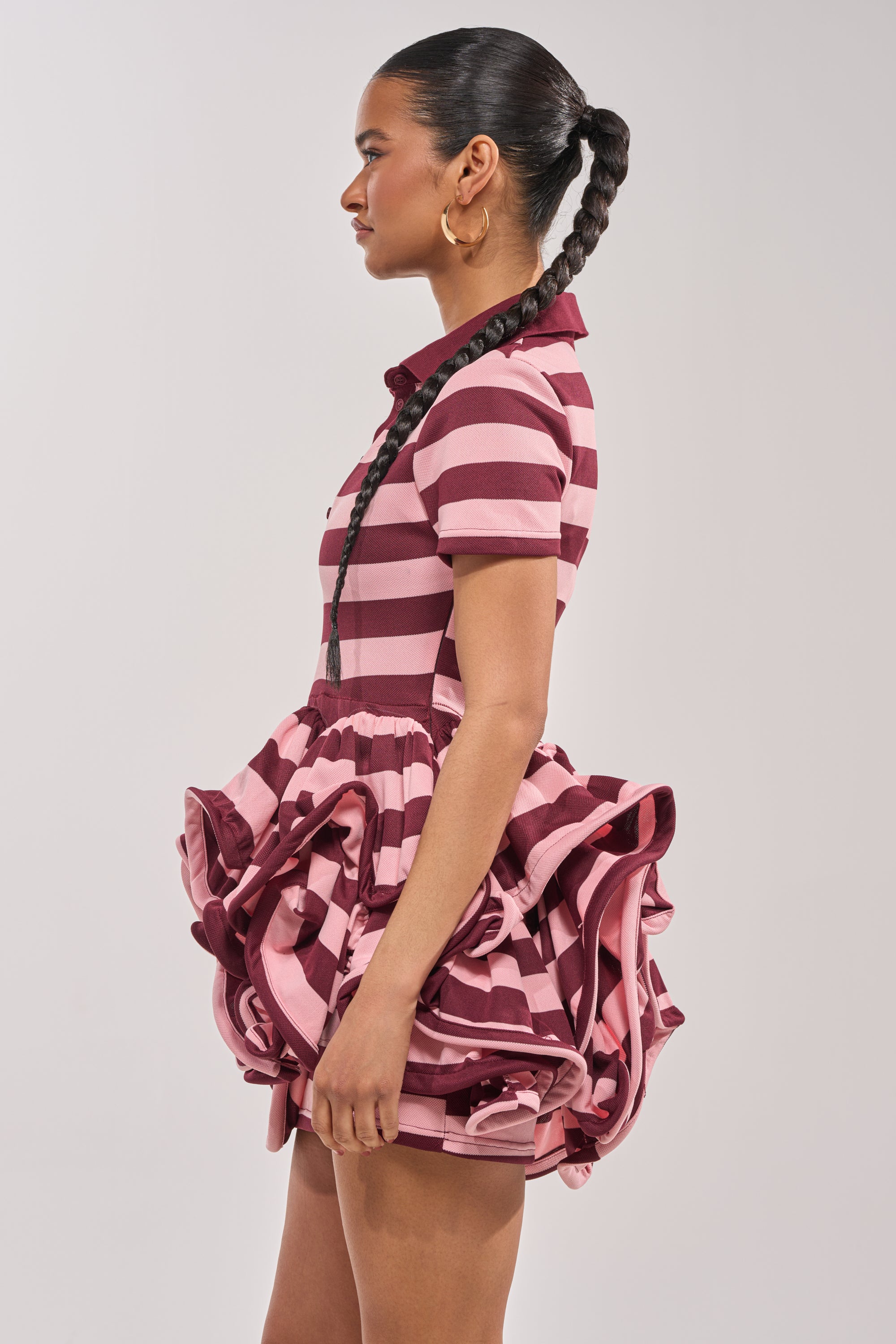 A woman stands in profile, wearing the OFFICE SIREN STRIPED SHIRT DRESS IN BURGUNDY, featuring pink and burgundy stripes and a ruffled mini skirt. She sports a long braid and large hoop earrings against a plain background. 2