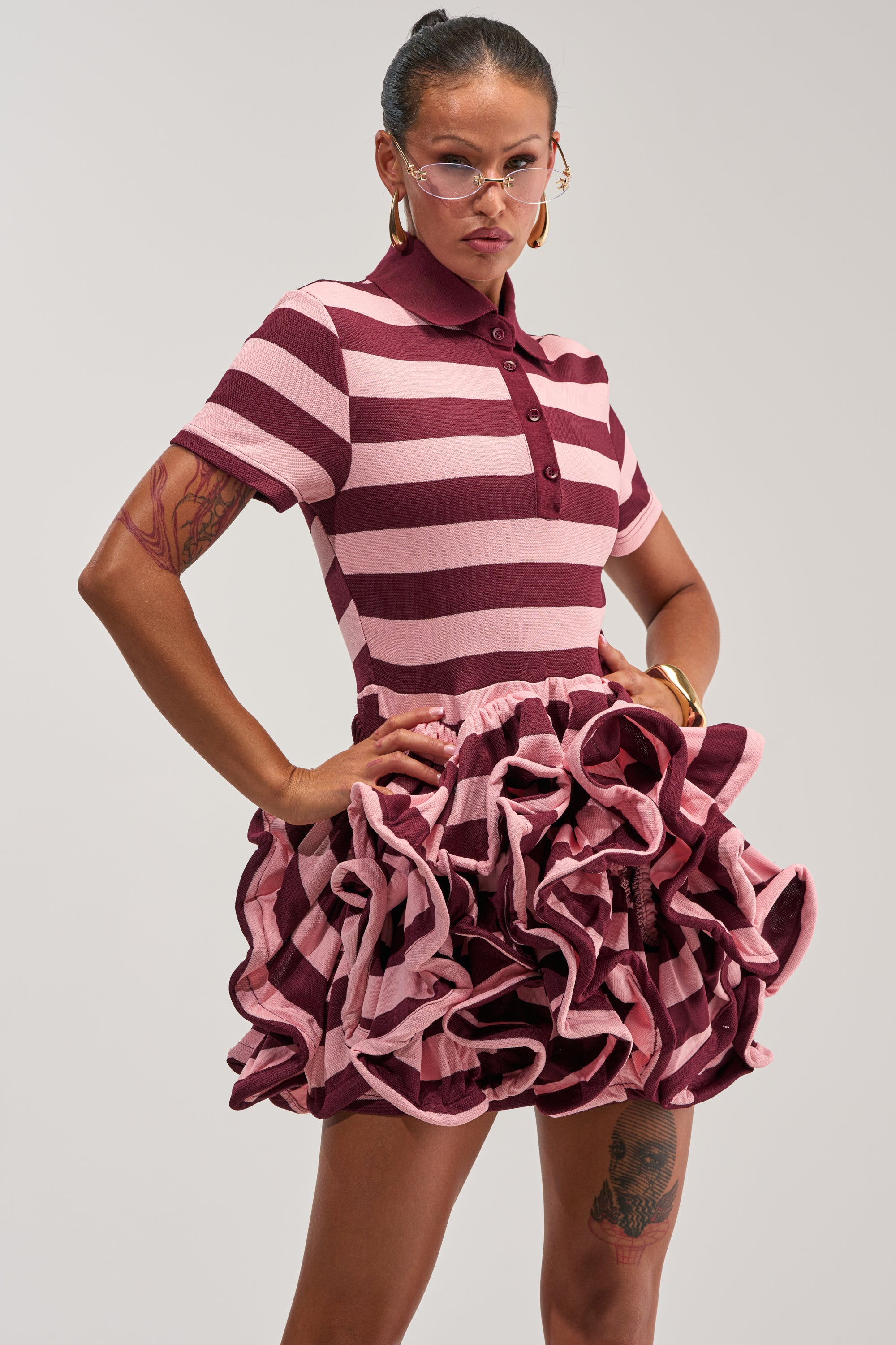 A person wearing glasses, hoop earrings, and the OFFICE SIREN STRIPED SHIRT DRESS IN BURGUNDY—a pink and burgundy striped mini dress with a ruffled skirt—stands confidently with hands on hips against a plain background.