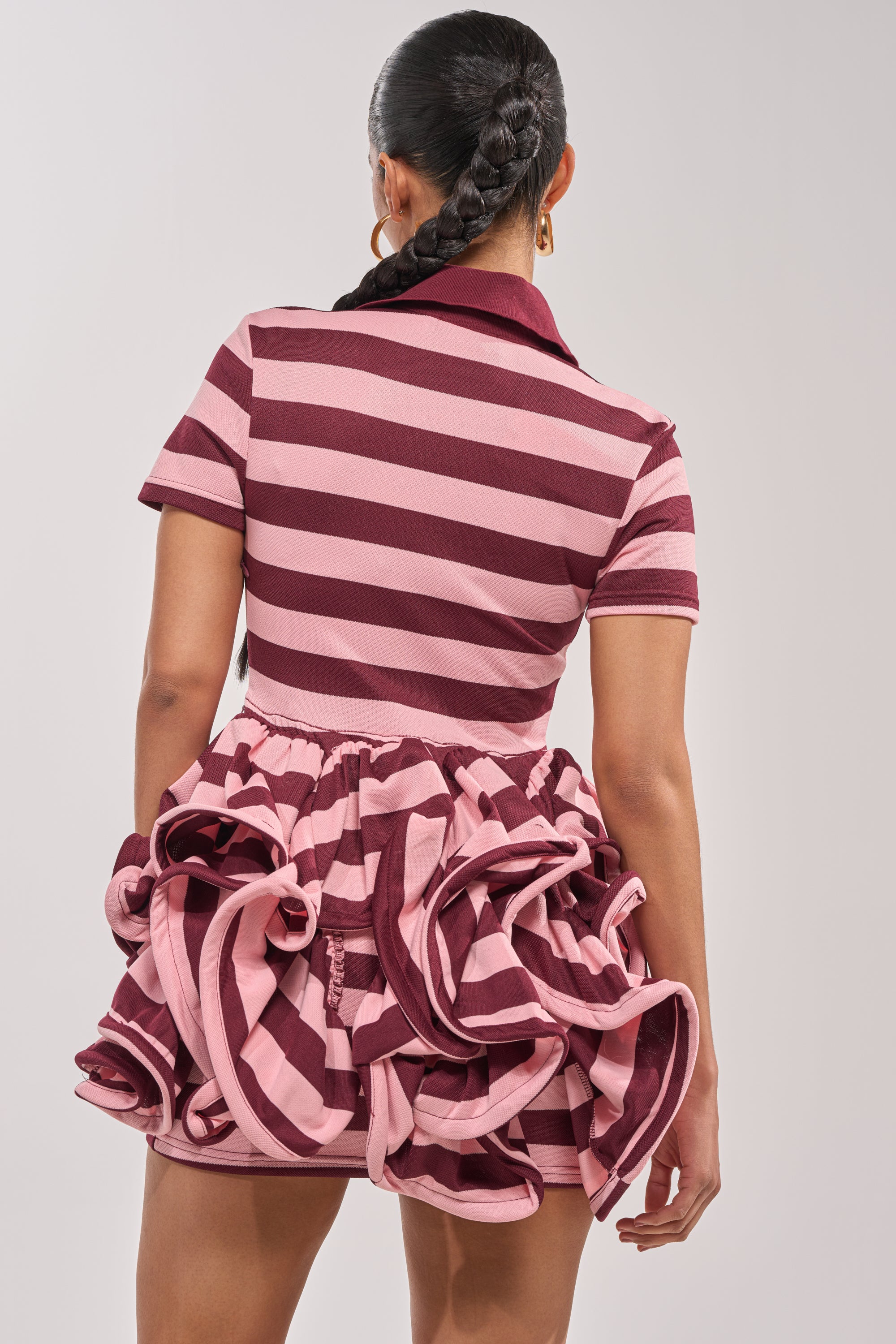 A person with a long braided ponytail is seen from the back wearing the OFFICE SIREN STRIPED SHIRT DRESS IN BURGUNDY, featuring maroon and pink stripes, a ruffle mini skirt, and set against a plain light background. 3