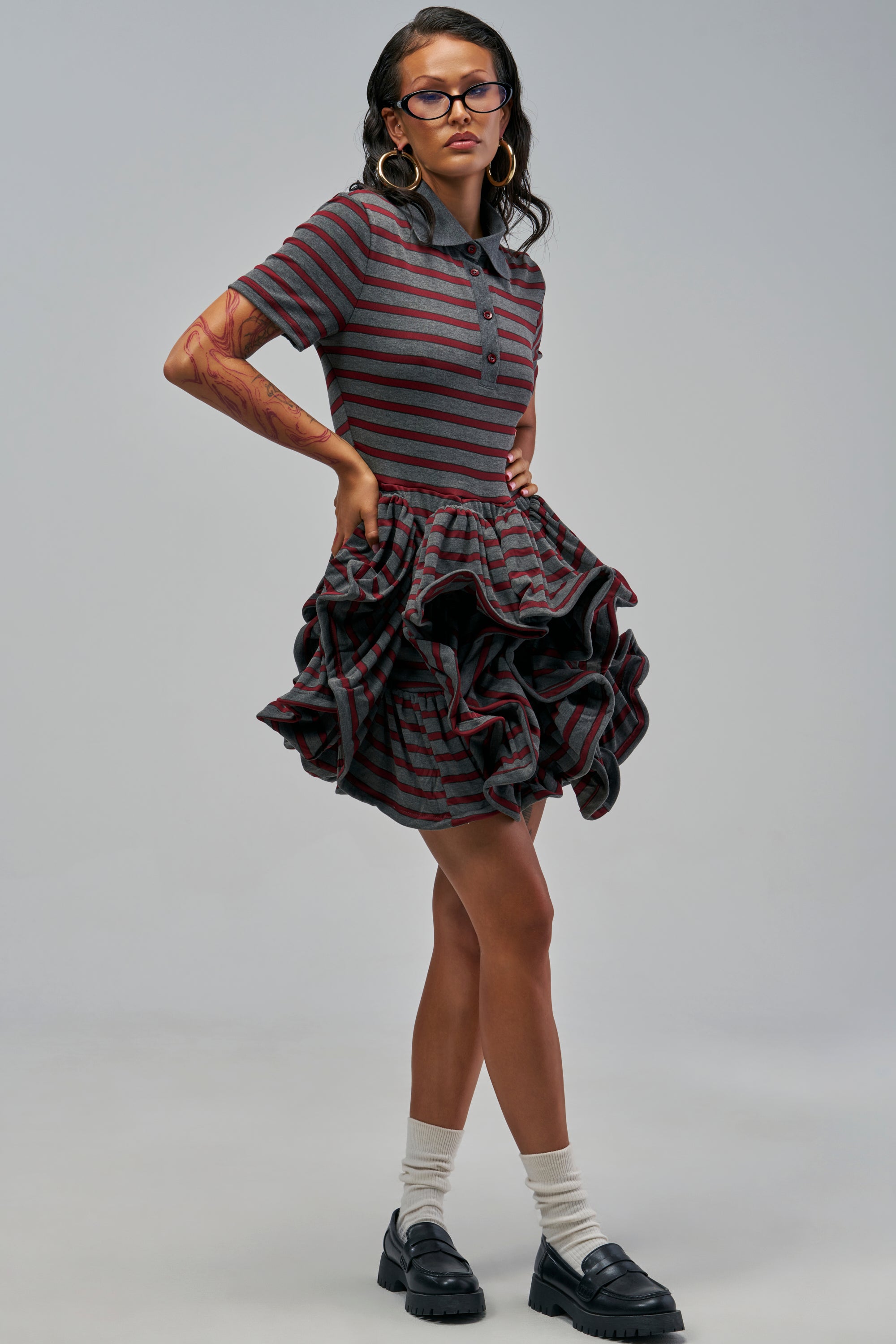 A woman wearing the SHE’S TOO COOL T-SHIRT DRESS IN GREY MULTI, featuring a grey and maroon striped polo top with a voluminous ruffled skirt, white socks, and black loafers.