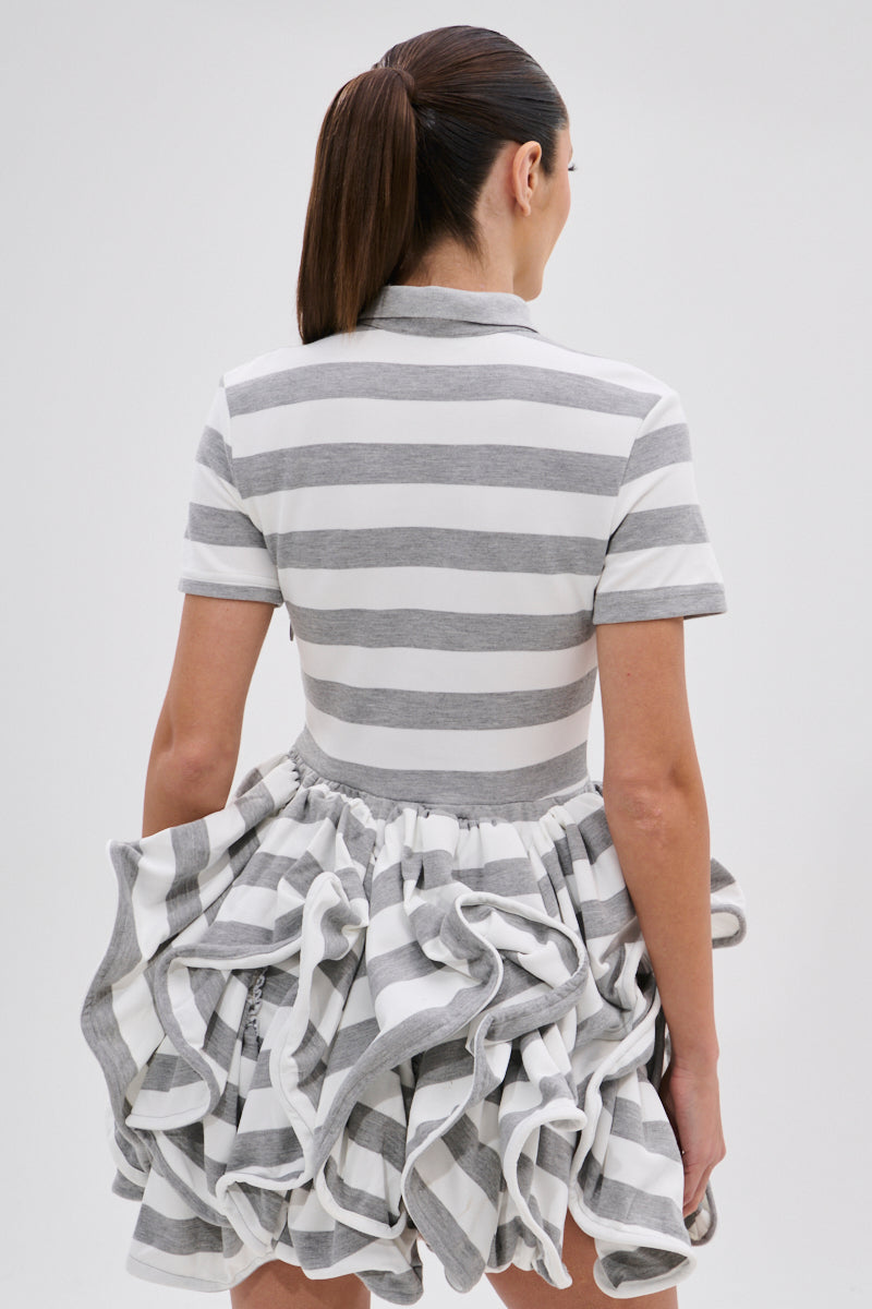 A woman with long brown hair in a ponytail wears the OFFICE SIREN STRIPED SHIRT DRESS, featuring a fitted knit top and ruffled bubble skirt in gray-and-white stripes. She stands with her back to the camera against a plain background.