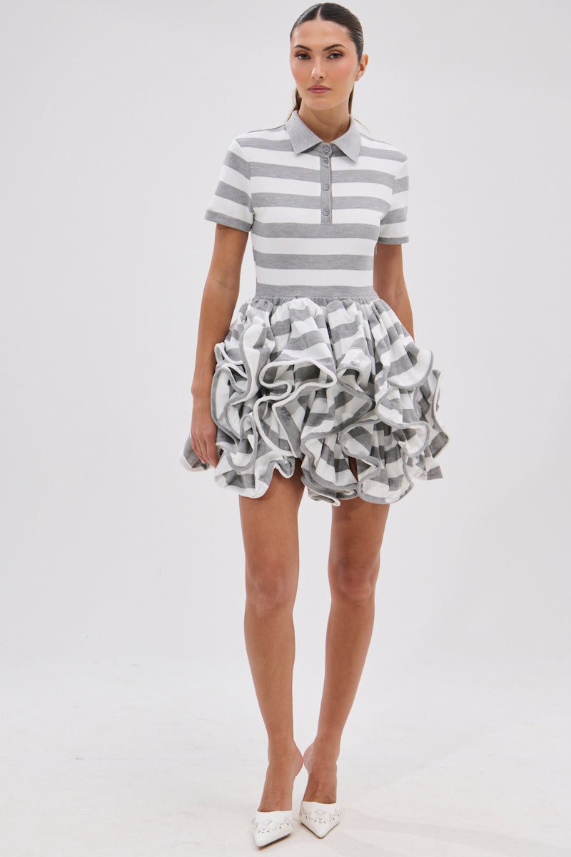 A woman stands against a plain background wearing the OFFICE SIREN STRIPED SHIRT DRESS in grey and white stripes. Her hair is pulled back, and she completes the look with white pointed-toe heels.