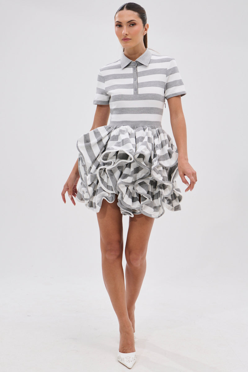 A woman models the OFFICE SIREN STRIPED SHIRT DRESS, featuring grey and white stripes, a polo collar, and a ruffled bubble skirt. She pairs it with white pointed-toe heels against a plain white background.