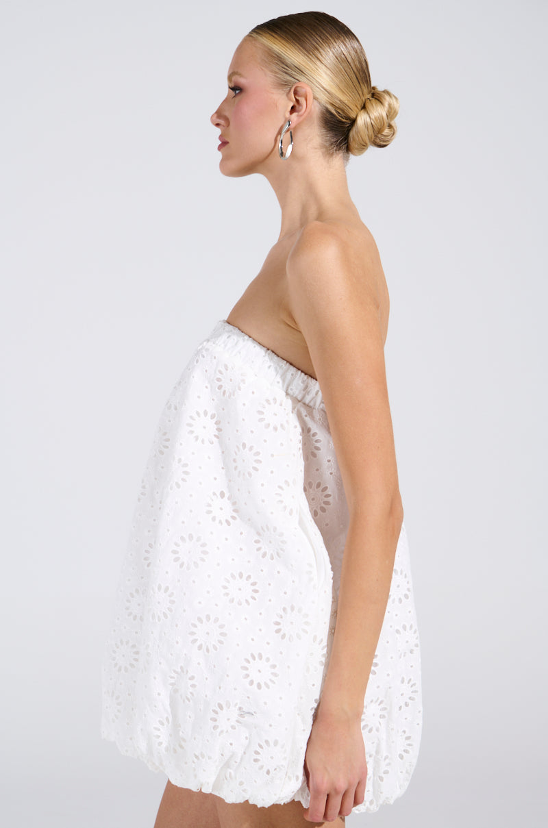 A person with slicked-back hair and silver hoop earrings models a strapless silhouette, the ALL FOR LOVE BUBBLE MINI DRESS in white eyelet-pattern, standing sideways against a plain background.