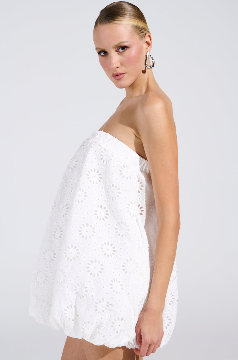 Modeling the ALL FOR LOVE BUBBLE MINI DRESS, a woman showcases its strapless silhouette and textured floral pattern. Her hair is slicked back with hoop earrings, and she poses against a plain light background, elegantly looking over her shoulder.