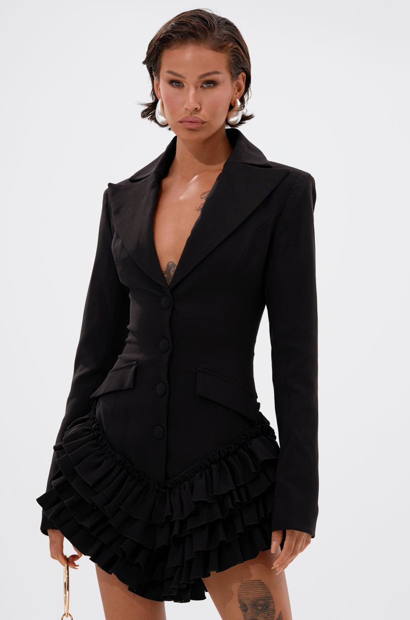 A woman stands against a plain light background wearing the STRICTLY BUSINESS BLAZER ROMPER IN BLACK, featuring a chic deep neckline, ruffled layered skirt, and button details. She has short hair, hoop earrings, and tattoos on her thigh and arm.