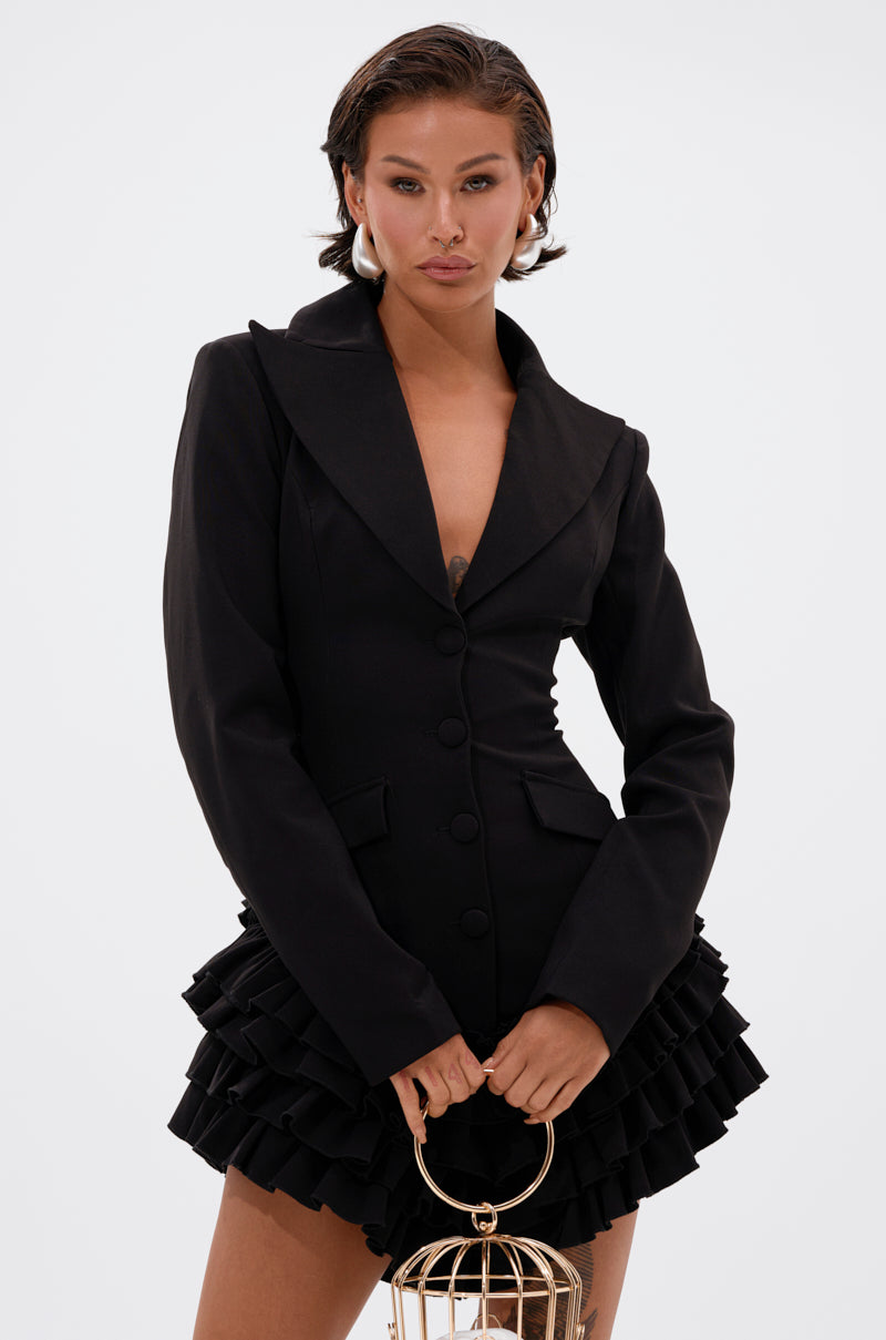 A woman poses confidently in the STRICTLY BUSINESS BLAZER ROMPER IN BLACK, featuring a deep V-neck and ruffled skirt. She holds a gold circular handbag, with slicked-back hair and bold makeup against a plain white background.