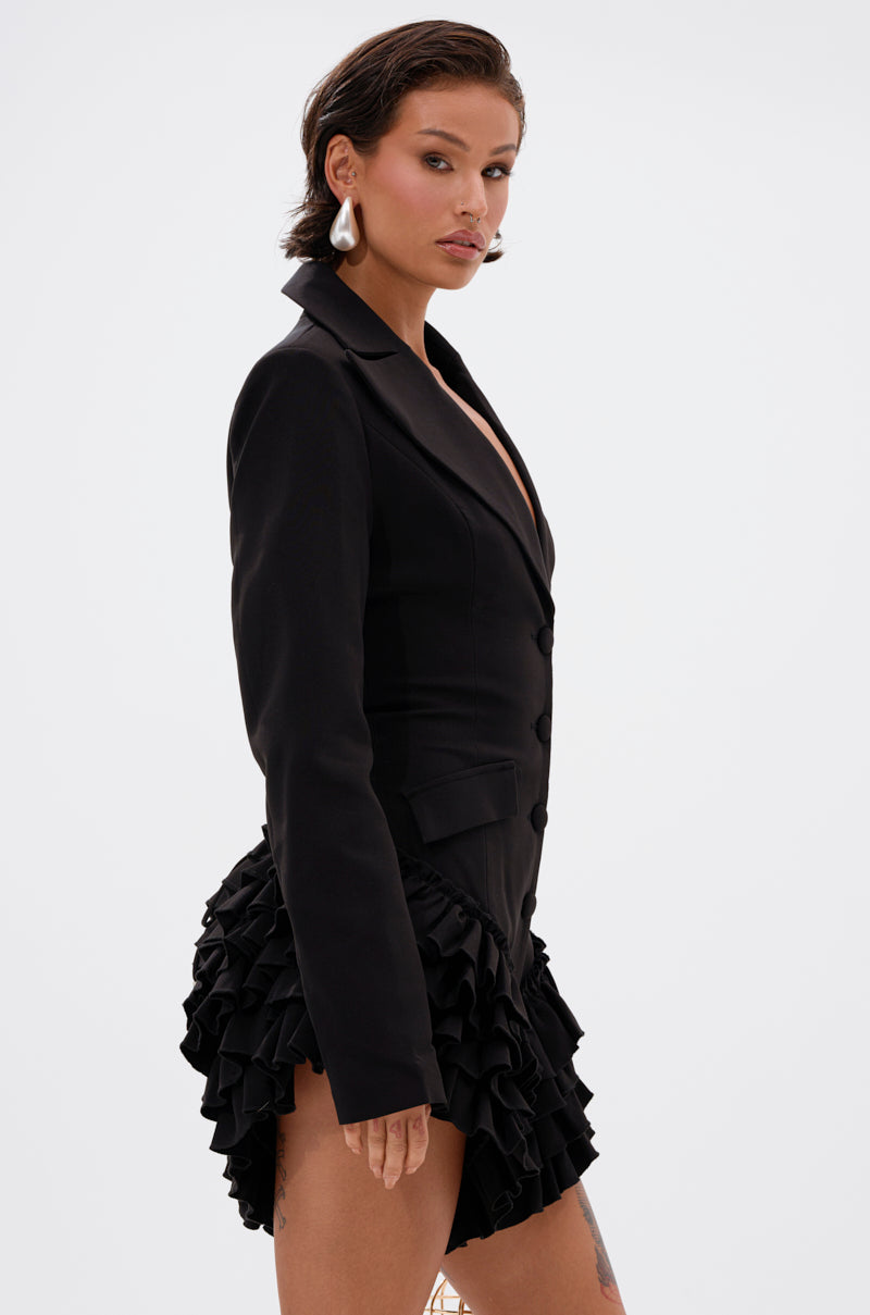 A woman with short hair and statement earrings poses confidently in the STRICTLY BUSINESS BLAZER ROMPER IN BLACK, featuring a fitted silhouette and tiered ruffle layers at the bottom, against a plain white background.