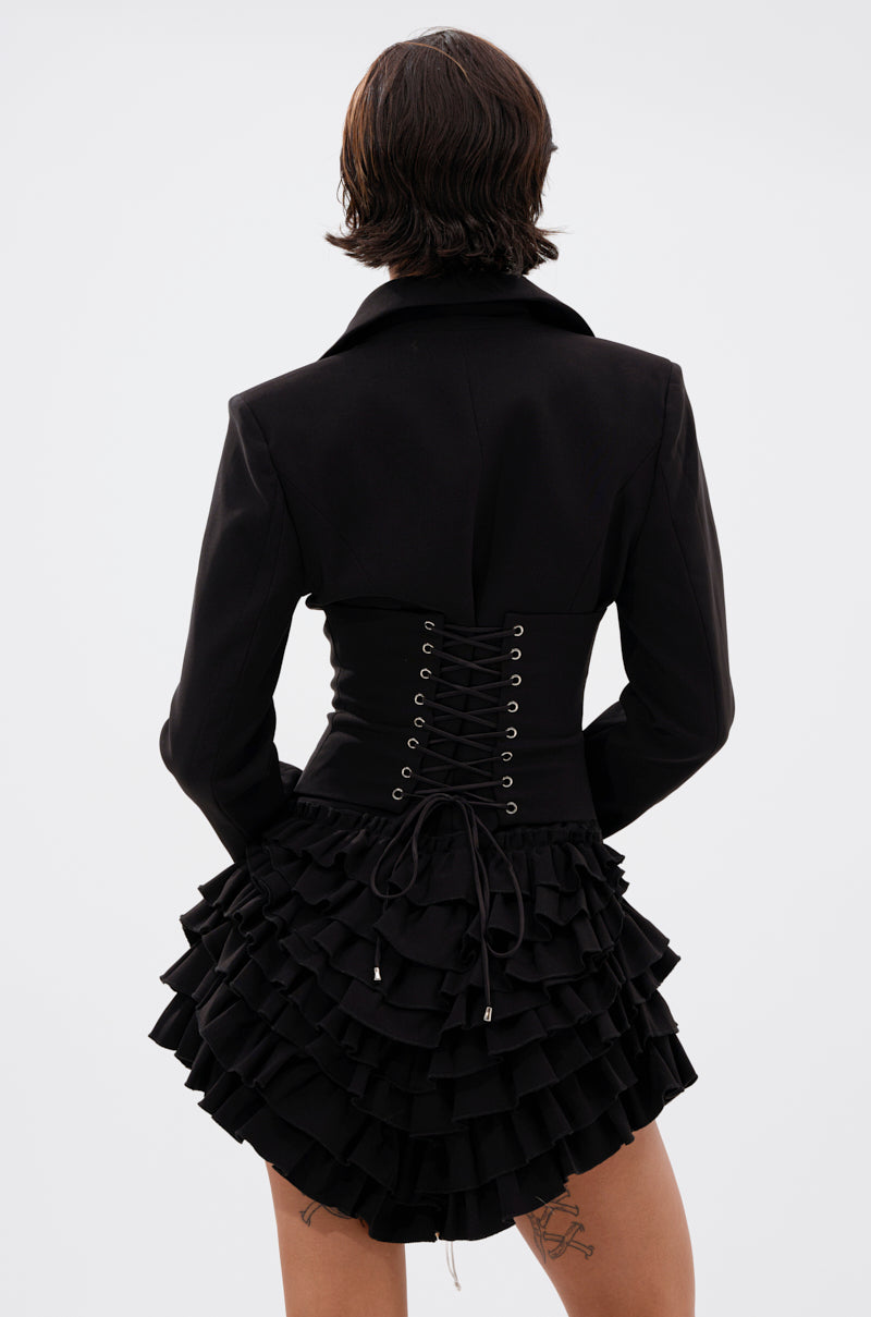 A person with short brown hair, seen from behind, wears the STRICTLY BUSINESS BLAZER ROMPER IN BLACK featuring a lace-up corset back and layered ruffled skirt. Tattoos are visible on their thighs against a plain light background.