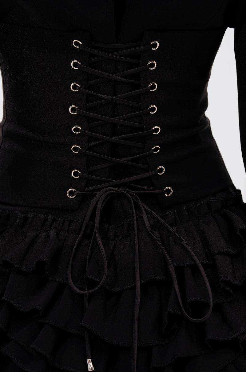 Close-up of the STRICTLY BUSINESS BLAZER ROMPER IN BLACK, styled with a black corset featuring silver eyelets and lace-up cords tied in a bow at the waist, worn over a ruffled skirt. The matte fabric enhances its sleek, tailored look.