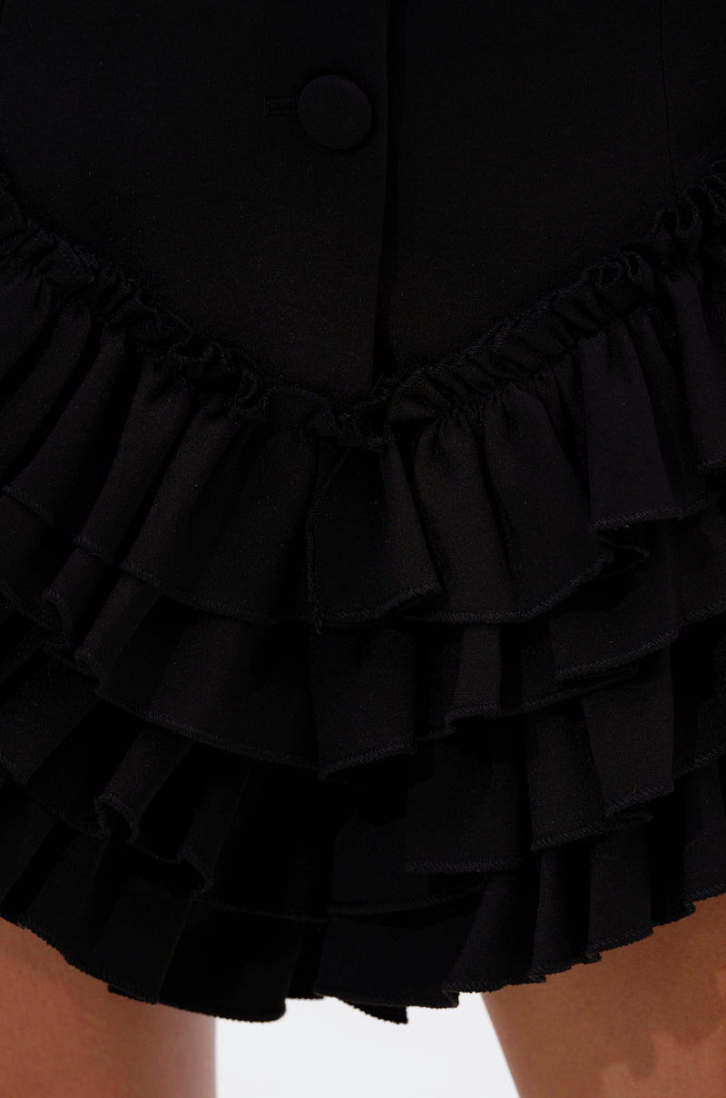 Close-up of the STRICTLY BUSINESS BLAZER ROMPER IN BLACK, featuring layered, pleated ruffles and a top button. This sleek piece ends just above the knees, showcasing a bold, modern style.