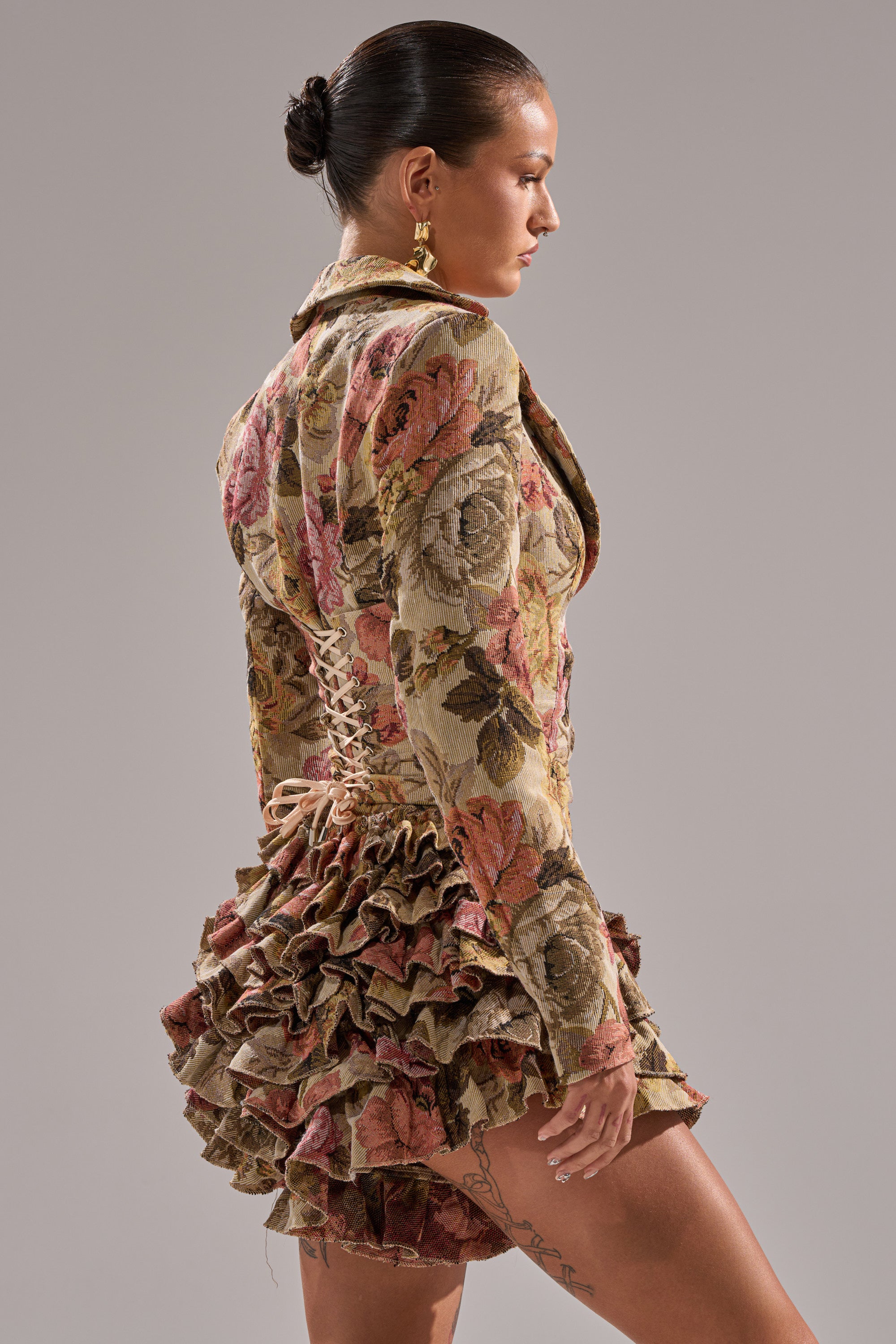 A woman with slicked-back hair wears the FLORAL HEAVEN TAPESTRY MINI DRESS, which features a fitted bodice and ruffle skirt. She stands in profile against a plain, neutral background.