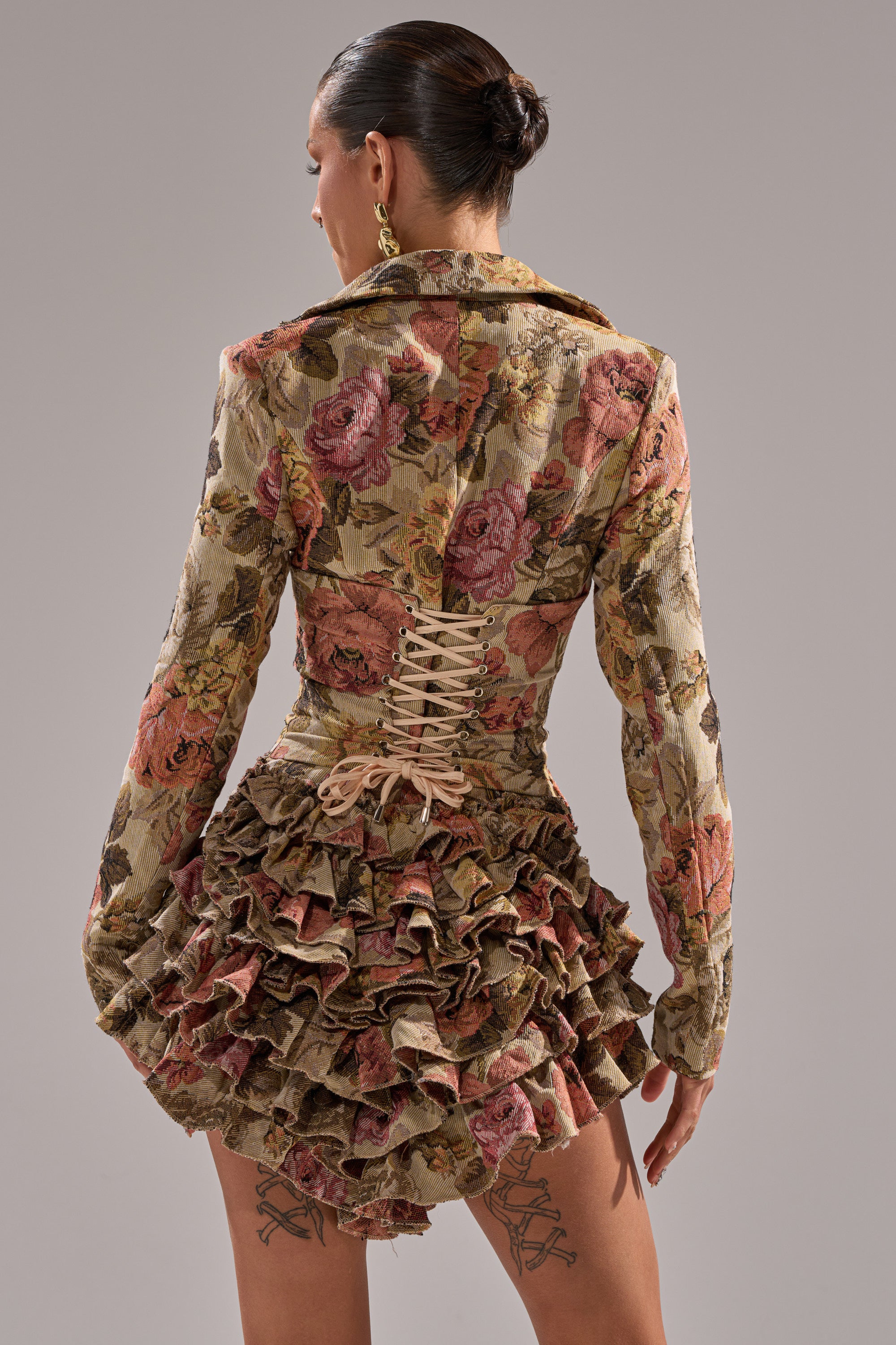 A person stands with their back to the camera, showcasing the FLORAL HEAVEN TAPESTRY MINI DRESS featuring a fitted, laced-up waist and layered ruffle skirt. Their hair is in a sleek bun and leg tattoos are visible.