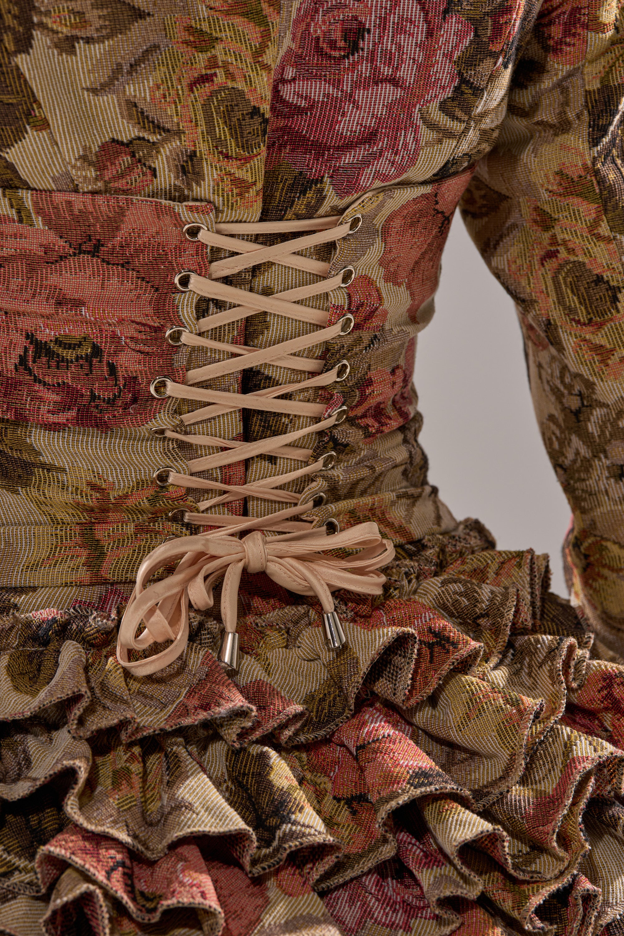 Close-up of the back of the FLORAL HEAVEN TAPESTRY MINI DRESS, a floral jacquard mini in brown, pink, and green with a beige lace-up corset detail and waist ruffles.