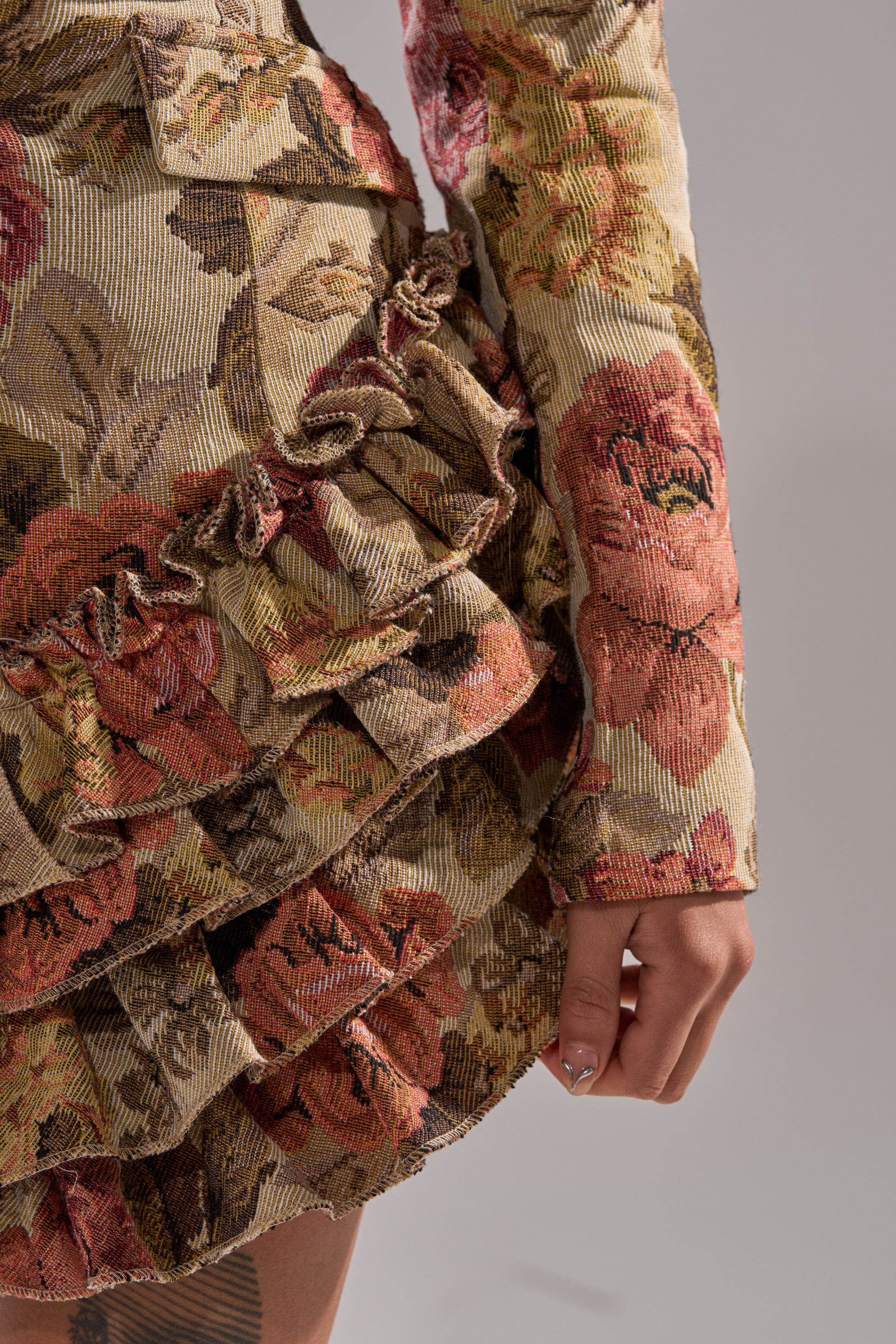 Close-up of a person wearing the FLORAL HEAVEN TAPESTRY MINI DRESS, a floral jacquard mini dress with layered ruffle skirt and long sleeves in warm earthy tones. Their hand rests by their side, revealing a manicure and part of a thigh tattoo.